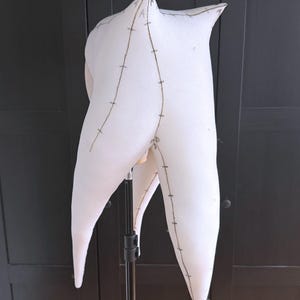 K&ouml;nnte beinhalten: Eine wei&szlig;e, abstrakte Stoffform mit spitzen Kanten und geschwungenen Linien. Die Form wird von einem Metallst&auml;nder getragen. Braune N&auml;hte umrei&szlig;en die Form und f&uuml;gen dem weichen, strukturierten Material Details hinzu. Das Design deutet auf ein skulpturales oder k&uuml;nstlerisches St&uuml;ck hin.