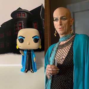 Peut inclure: Figurine Funko Pop d'un personnage avec des fards à paupières bleus, des boucles d'oreilles jaunes et une robe bleue. La figurine est devant une image en noir et blanc d'une maison.
