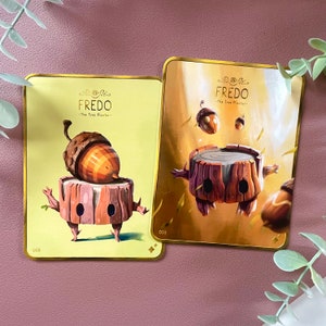 May include: Two illustrated cards with a gold background and gold trim. The cards feature a cartoon character called "Fredo - The Tree Planter". The first card shows Fredo with a large acorn on his head. The second card shows Fredo floating in the air with smaller acorns around him.