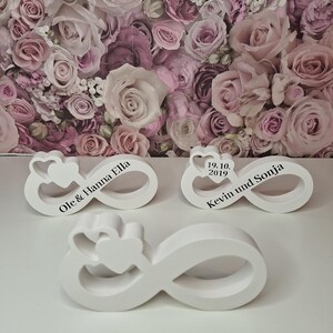 Könnte beinhalten: Drei weiße Holz-Unendlichkeits-Symbole mit gravierten Namen und Daten. Die Ornamente befinden sich auf einem Hintergrund aus rosa Rosen.