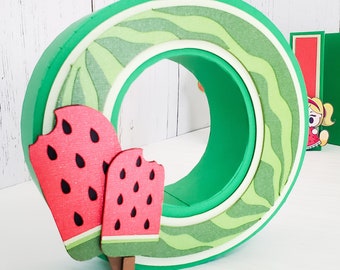 Könnte beinhalten: Ein grüner, wassermelonenförmiger Buchstabe O mit zwei Wassermelonen-Eis am Rand. Die Eis am Stiel sind rot mit schwarzen Kernen und einem grünen Stiel.