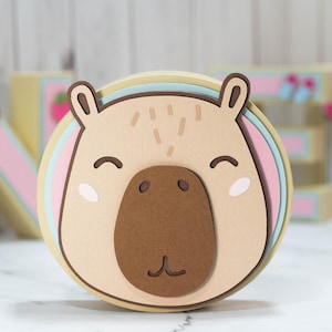 May include: A layered, circular craft featuring a capybara face. The design includes shades of tan, brown, pink, and blue. The capybara has a large brown nose, small eyes, and a smiling mouth. The ears are rounded and brown.