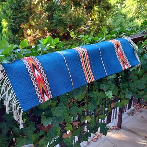 May include: A blue woven table runner with a geometric pattern in red, yellow, and white. The runner has white fringe on the ends and is displayed outdoors. The runner is approximately 30 cm wide and 122 cm long.