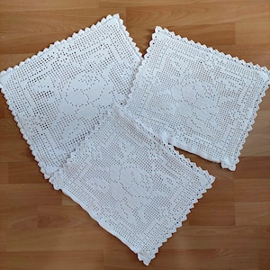 May include: Three white, square crochet doilies with a floral design. The doilies are arranged on a wooden surface. Each doily has a scalloped edge. The crochet work is detailed and intricate, showcasing a classic, handmade aesthetic.