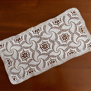 Puede incluir: Camino de mesa blanco de ganchillo con un delicado patrón floral. El camino de mesa presenta un diseño repetitivo de espirales y flores.