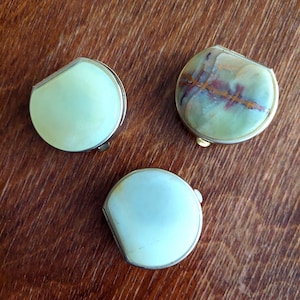 Op de afbeelding: Drie kleine, vintage compactpoeders met afgeronde, asymmetrische vormen. Twee hebben lichtgroene oppervlakken, terwijl de derde een gevlekt groen en bruin patroon heeft. Elke compactpoeder heeft een zilverkleurig metalen frame.