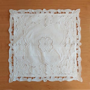 Peut inclure: Nappe carrée blanche brodée avec un motif floral. Le centre présente une fleur détaillée, entourée d'une forme de trèfle à quatre feuilles. Les bords sont festonnés et ornés de motifs floraux découpés. Cette pièce décorative est probablement utilisée pour la décoration intérieure.