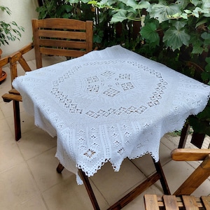 Könnte beinhalten: Weiße Häkeltischdecke mit einem zarten Blumenmuster, drapiert über einen Holztisch mit zwei Stühlen.