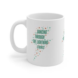 Könnte beinhalten: Weißer Keramikbecher mit gebogenem Henkel. Der Becher hat ein Blitzdesign, das mit türkisfarbenen und orangefarbenen Sternen gefüllt ist. Der Text "DANCING THROUGH THE LIGHTNING STRIKES" ist in Türkis gedruckt.
