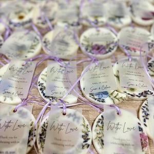 May include: A collection of round white wax ornaments embedded with dried flowers and lavender sprigs. Each ornament features a translucent rectangular tag tied with a purple ribbon, printed with the text With Love, Thank you for celebrating with us, APRIL 18, 2026, and Sam & Celine.