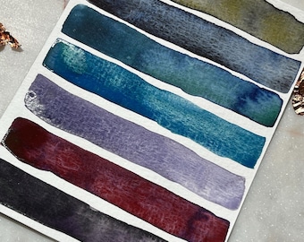 May include: A rectangular white paper card featuring six horizontal watercolor paint swatches arranged in a stack. The colors include olive green, dark charcoal, deep teal, bright turquoise, muted lavender, and a rich burgundy-purple gradient.