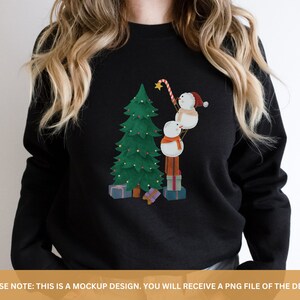 May include: A black sweatshirt with a Christmas-themed design featuring three snowmen building a Christmas tree. The snowmen are wearing red hats and the tree is decorated with green and red ornaments.
