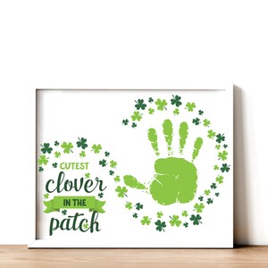 May include: A white frame with a green handprint surrounded by green shamrocks. The text "Cutest clover in the patch" is written in green.