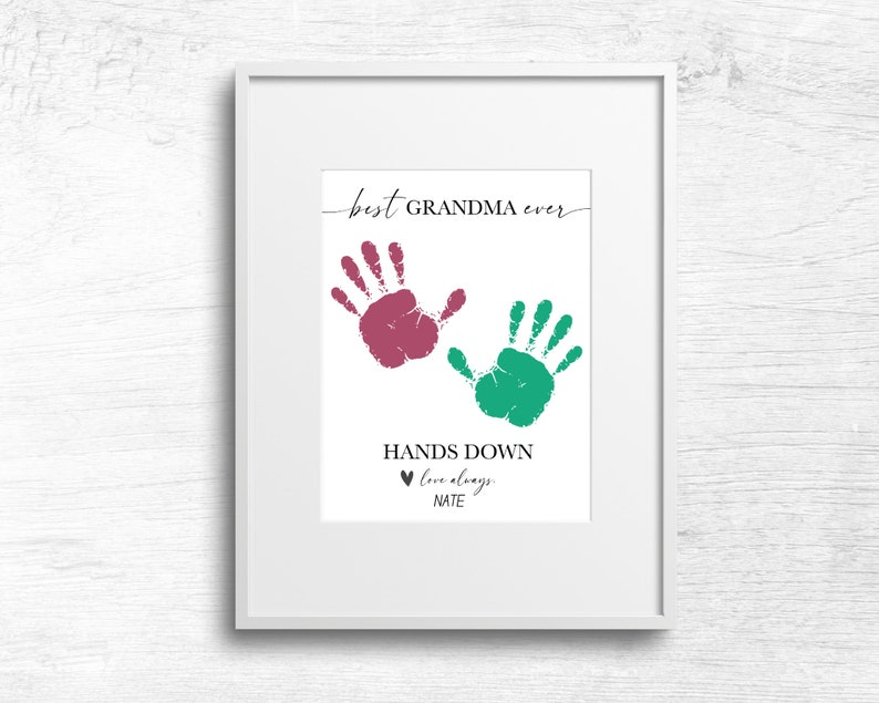 Op de afbeelding: Een wit ingelijst afdruk met twee handafdrukken, een roze en een groene, met de tekst "best GRANDMA ever" en "HANDS DOWN love always, NATE".