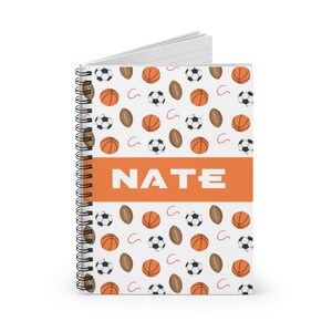 May include: A white spiral notebook with a colorful sports pattern. The cover features basketballs, footballs, and soccer balls. The name "NATE" is printed in orange on a band across the middle of the notebook.