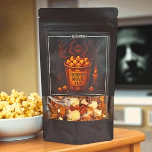 Könnte beinhalten: Ein schwarzer Beutel mit "Horror Movie Tea" mit einem transparenten Fenster, das die Teemischung zeigt. Das Etikett zeigt einen Popcorn-Eimer mit einem Auge und einer Kerze. Links steht eine Schüssel Popcorn, im Hintergrund ein verschwommener Fernsehbildschirm.