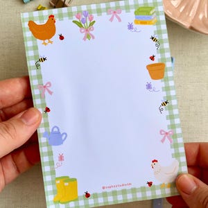 May include: A rectangular notepad featuring a white centre and a light green gingham border. The border is decorated with illustrations of a chicken, a white hen, yellow rain boots, a blue watering can, flowers, bows, bees, and ladybugs.