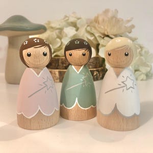 May include: Three wooden doll figures with painted faces and floral headbands. Each doll is wearing a different colored dress with a star and wand design. The dresses are pink, green, and white.