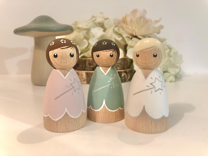 May include: Three wooden doll figures with painted faces and floral headbands. Each doll is wearing a different colored dress with a star and wand design. The dresses are pink, green, and white.