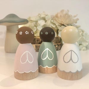 May include: Three wooden peg dolls with painted faces and floral details. The dolls are wearing dresses in pink, green, and white with a heart design. The dolls are standing on a wooden base.