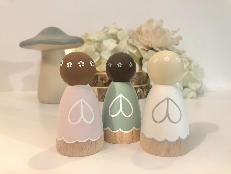 May include: Three wooden peg dolls with painted faces and floral details. The dolls are wearing dresses in pink, green, and white with a heart design. The dolls are standing on a wooden base.