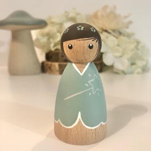 May include: A wooden doll with brown hair and a light blue dress. The doll is holding a white wand with a star on the end. The doll is painted with a whimsical style.