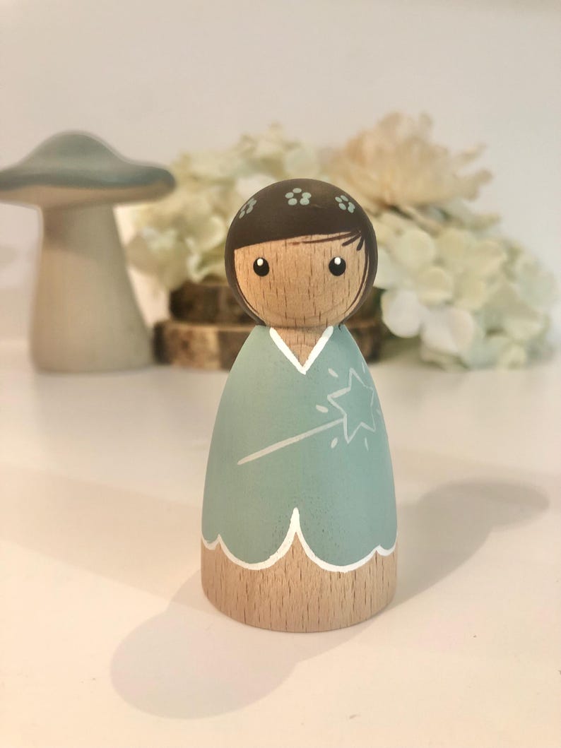 May include: A wooden doll with brown hair and a light blue dress. The doll is holding a white wand with a star on the end. The doll is painted with a whimsical style.