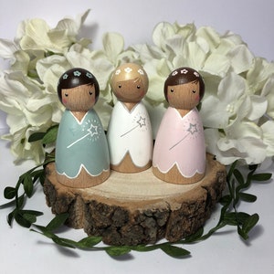 May include: Three wooden peg dolls dressed as fairies. Each doll has a different colored dress: light blue, white, and pink. The dolls have brown hair and wear flower crowns. They are standing on a wooden slice.