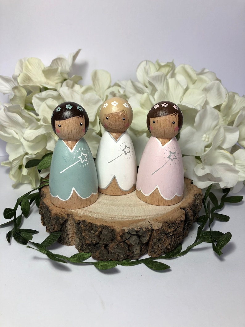 May include: Three wooden peg dolls dressed as fairies. Each doll has a different colored dress: light blue, white, and pink. The dolls have brown hair and wear flower crowns. They are standing on a wooden slice.