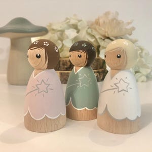 May include: Three wooden dolls with painted faces and floral headbands. Each doll is wearing a different colored dress with a white trim and a star design. The dolls are standing on a white surface.
