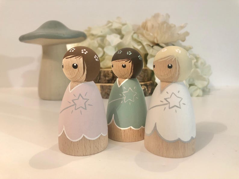 May include: Three wooden dolls with painted faces and floral headbands. Each doll is wearing a different colored dress with a white trim and a star design. The dolls are standing on a white surface.