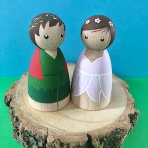 May include: Two wooden peg dolls, one with a green and red outfit and a leafy crown, the other with a pink dress and a white flower crown, standing on a wooden slice.