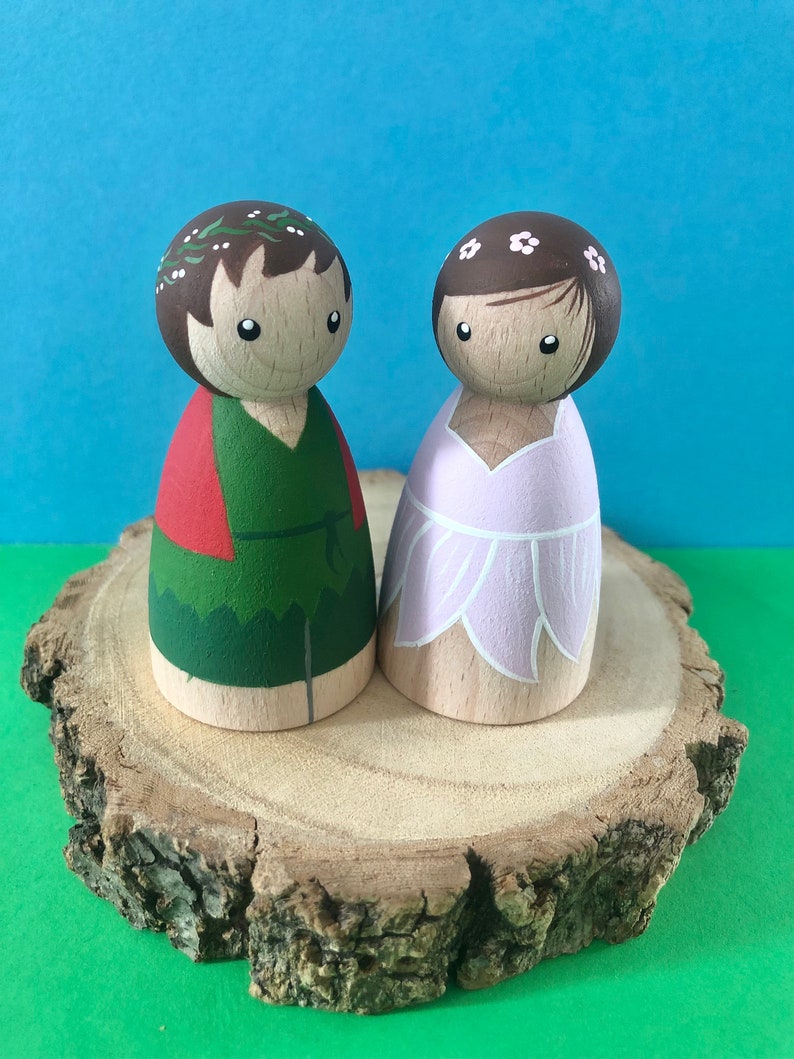 May include: Two wooden peg dolls, one with a green and red outfit and a leafy crown, the other with a pink dress and a white flower crown, standing on a wooden slice.