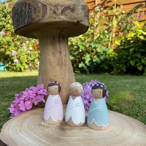 May include: Three wooden fairy dolls with painted faces and floral headbands. Each doll is wearing a different colored dress with a white star and wand. The dolls are standing on a wooden slice.