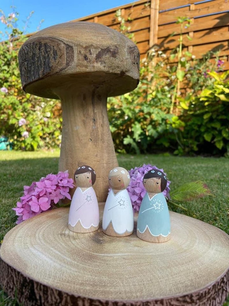 May include: Three wooden fairy dolls with painted faces and floral headbands. Each doll is wearing a different colored dress with a white star and wand. The dolls are standing on a wooden slice.