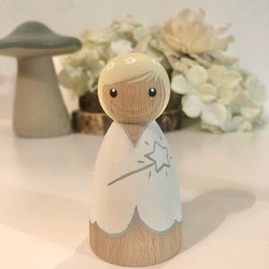May include: A wooden doll with blonde hair and a white dress. The doll is holding a wand with a star on the end. The doll is painted with a light blue and gray color scheme.
