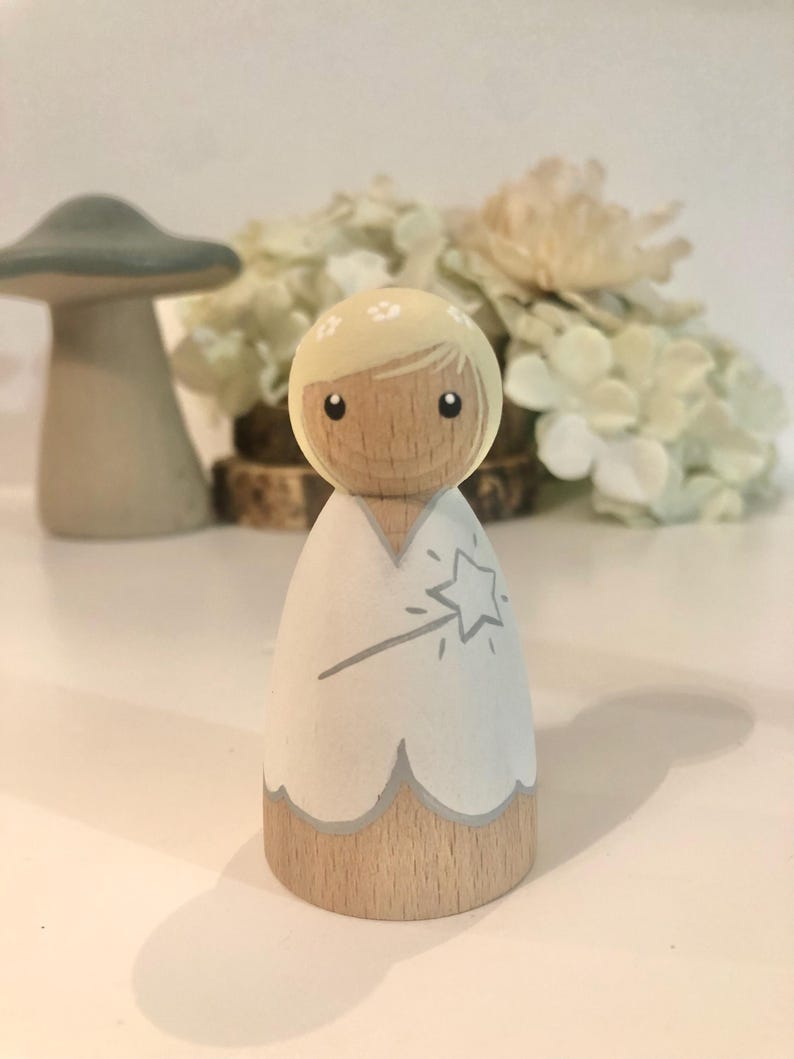 May include: A wooden doll with blonde hair and a white dress. The doll is holding a wand with a star on the end. The doll is painted with a light blue and gray color scheme.