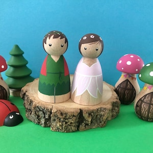 May include: A set of wooden peg dolls dressed as a fairy and a forest creature. The dolls are standing on a wooden slice with mushroom houses and a ladybug.