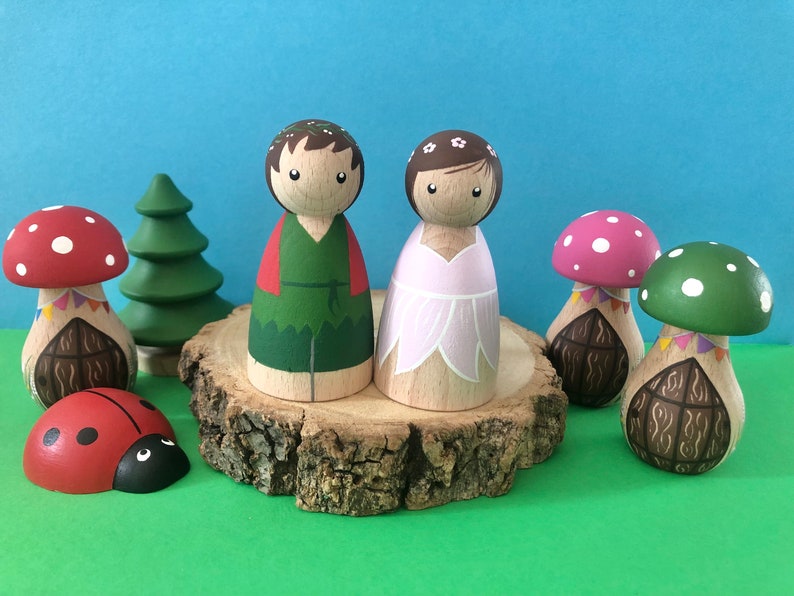 May include: A set of wooden peg dolls dressed as a fairy and a forest creature. The dolls are standing on a wooden slice with mushroom houses and a ladybug.