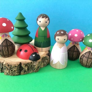 May include: A set of wooden dollhouse figures, including a girl and boy doll, a ladybug, and three mushroom houses with doors and flags. The figures are painted in bright colours and the houses have a whimsical design.