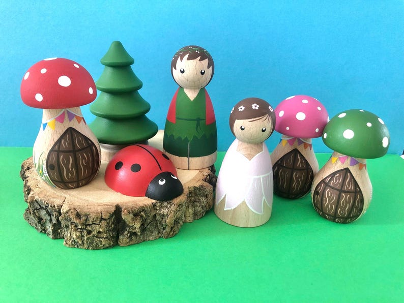 May include: A set of wooden dollhouse figures, including a girl and boy doll, a ladybug, and three mushroom houses with doors and flags. The figures are painted in bright colours and the houses have a whimsical design.