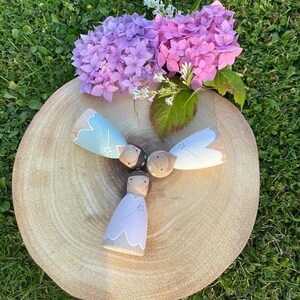 May include: Three wooden dolls with painted faces and star details. The dolls are wearing pastel blue, light purple, and white dresses. The dolls are arranged in a circle on a wooden surface. The background is a green grassy area with pink flowers.