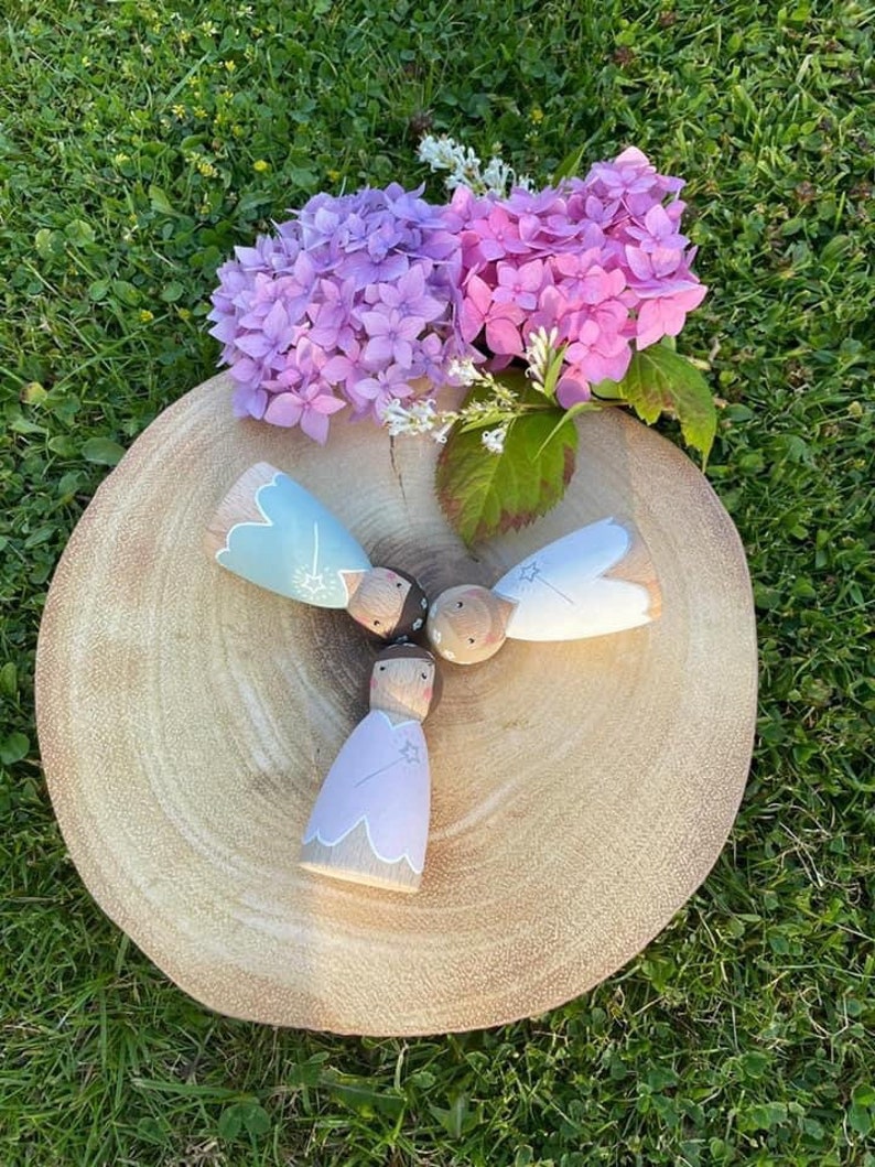 May include: Three wooden dolls with painted faces and star details. The dolls are wearing pastel blue, light purple, and white dresses. The dolls are arranged in a circle on a wooden surface. The background is a green grassy area with pink flowers.