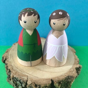 May include: Two wooden peg dolls, one dressed in green and red with a leafy crown, the other in a pink dress with a white flower crown, standing on a wooden slice.
