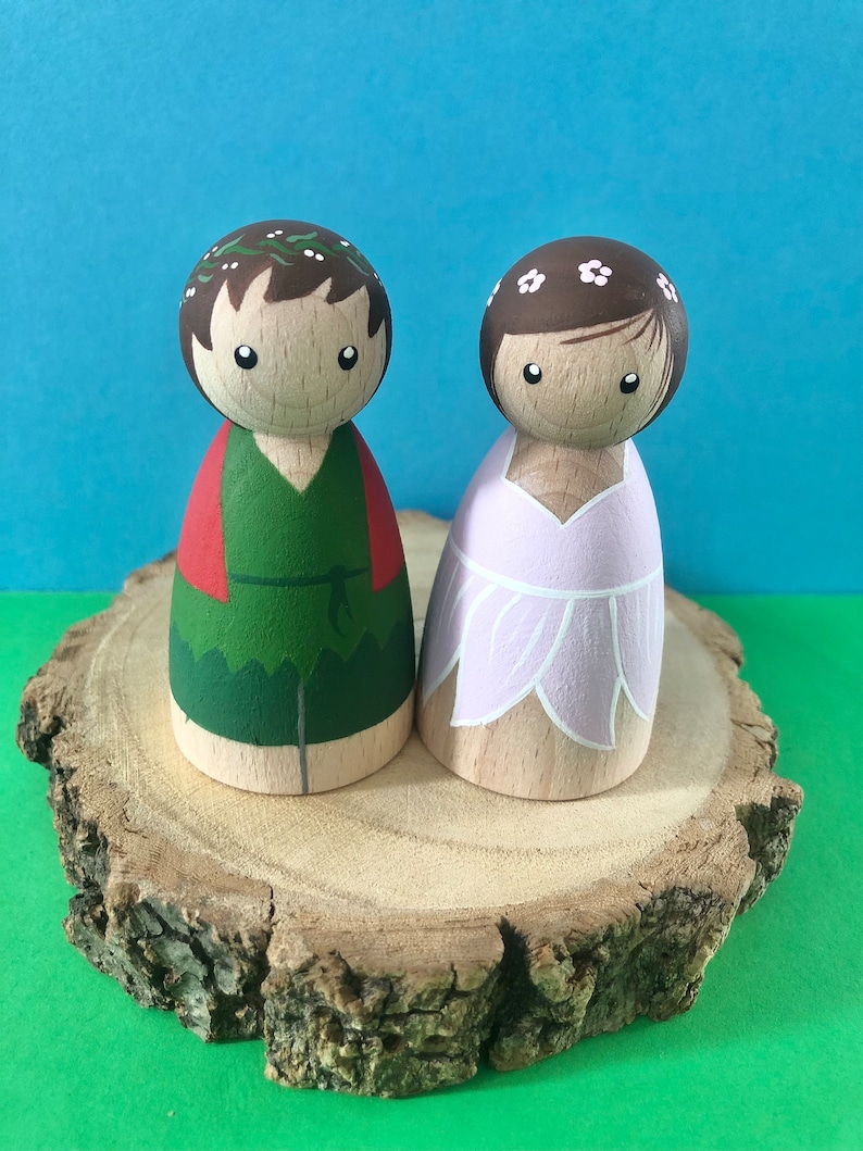 May include: Two wooden peg dolls, one dressed in green and red with a leafy crown, the other in a pink dress with a white flower crown, standing on a wooden slice.