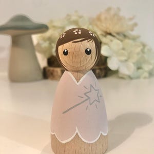 May include: A wooden doll with brown hair and a pink dress. The doll is holding a wand with a star on the end. The doll is painted with a whimsical style.