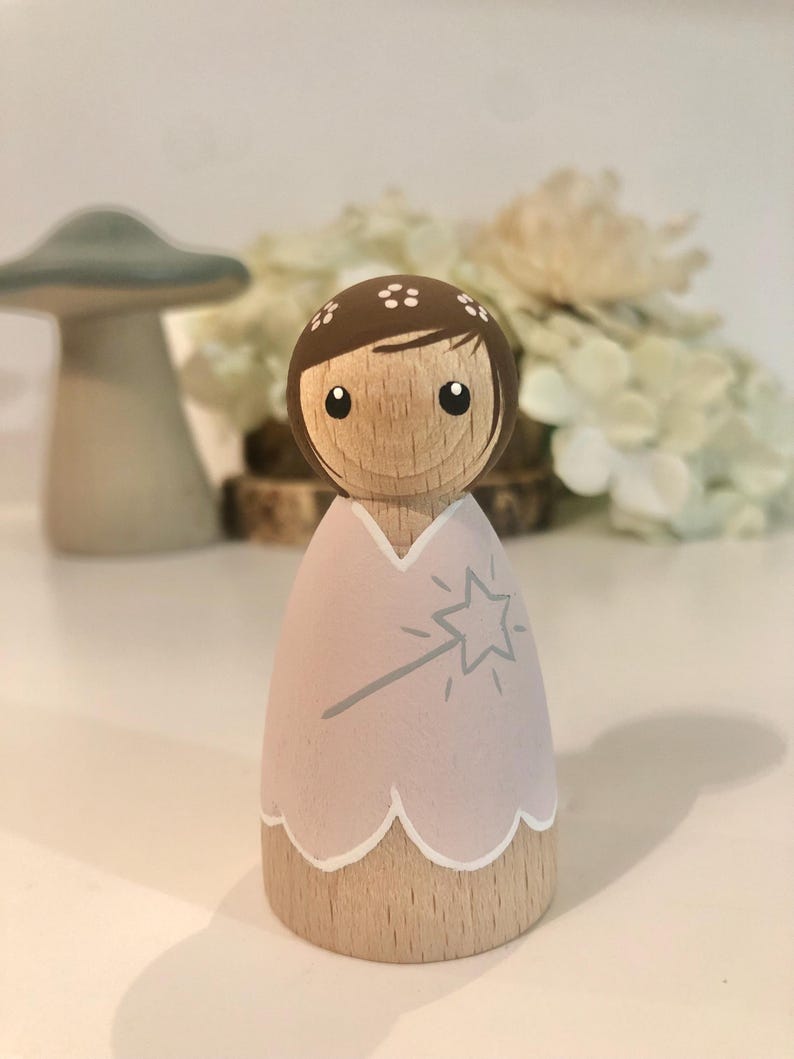 May include: A wooden doll with brown hair and a pink dress. The doll is holding a wand with a star on the end. The doll is painted with a whimsical style.