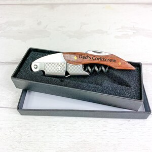 May include: A wooden and stainless steel corkscrew with the text "Dad's Corkscrew" engraved on the handle. The corkscrew is presented in a black gift box with a black interior.