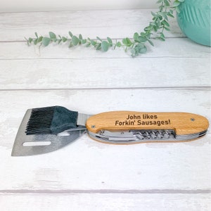 May include: A multi-tool with a wooden handle, featuring a spatula, bottle opener, and a brush. The handle is engraved with the text "John likes Forkin' Sausages!"