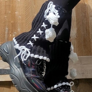 Puede incluir: Botas de plataforma negras con acabado brillante, combinadas con calentadores de piernas de punto negro. Los calentadores de piernas presentan detalles de ganchillo blanco, incluyendo pompones y cordones cruzados. Las botas tienen una suela gruesa.